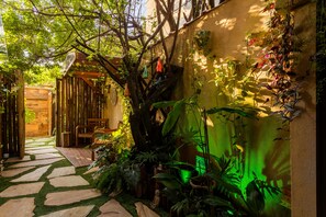 Un sentier de jardin mène à une structure en bois surmontée d’un banc, le tout étant entouré d’une végétation luxuriante et de plantes en pot.