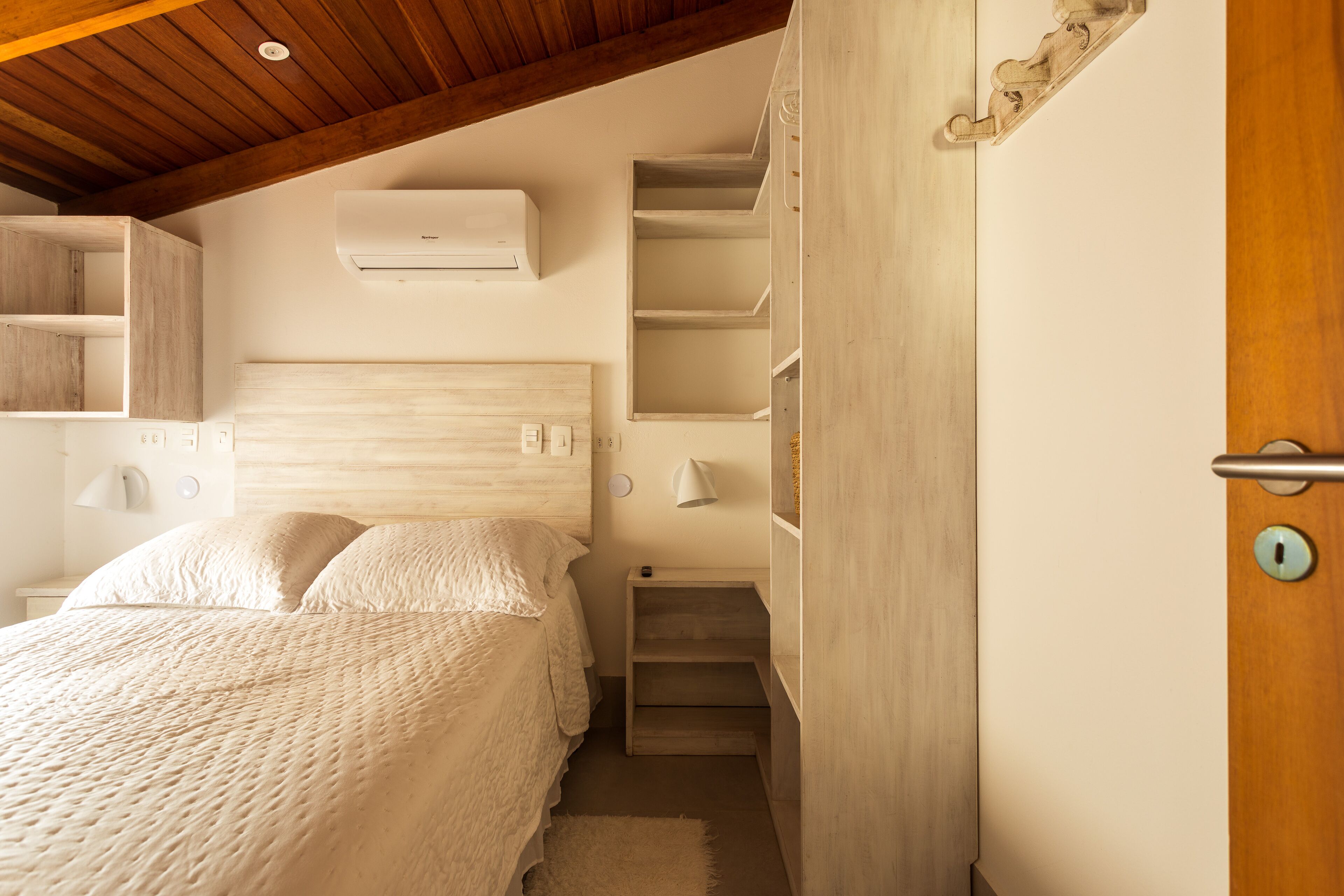 A bedroom with a wooden ceiling, a bed with a white bedspread, a nightstand, a wall-mounted air conditioner, and a door with a keyhole.