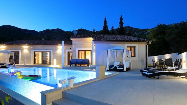 Area piscina con lettini, zona relax coperta e un edificio con più porte e finestre.