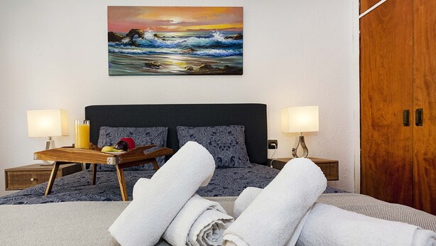 A neatly made bed with rolled white towels, a wooden bedside table with a fruit bowl, and a wall painting of a seascape.