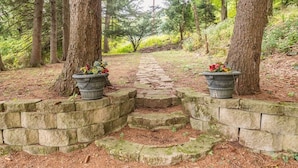 Ein Steinweg führt durch ein bewaldetes Gebiet, beidseitig jeweils mit je einem Pflanztopf.