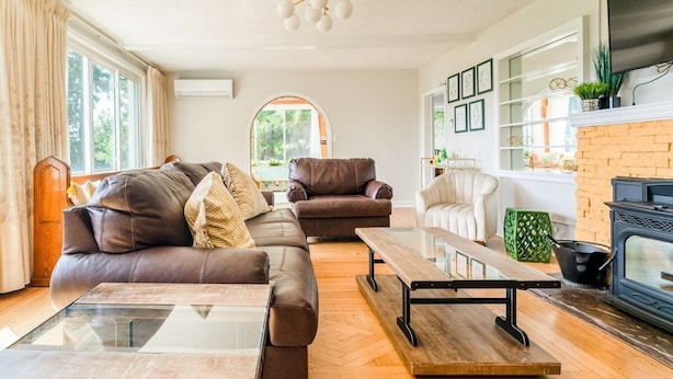 Ein Wohnzimmer mit braunem Ledersofa, einem Glastisch, einem Holznachttisch, einem Kamin und einem an der Wand montierten Fernseher.