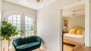 Ein Zimmer mit einem grünen Sofa, einem Holztisch, einem Bett mit gelben Kissen und Blick durch eine Tür.