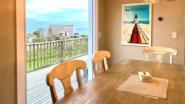 A dining area with a view of the sea, a poster of Nantucket, and a wooden table with chairs.