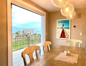 A dining area with a view of the ocean, a poster of Nantucket, and a wooden table with chairs. - Luxury Dreamy Madaket Destination - Beachside, Ocean Views & Lifelong Memories (Nantucket)