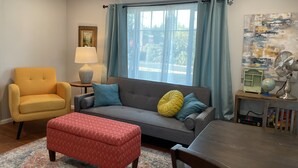 Ein Wohnzimmer mit einer Couch, einem Hocker, einem Sessel und einem Tisch mit einer Lampe.