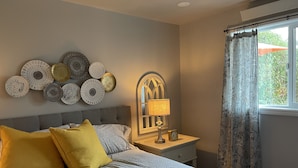Ein Schlafzimmer mit einem Bett, einem Nachttisch mit Lampe und Uhr, dekorativen Tellern an der Wand und einem Fenster mit Vorhängen.