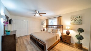 A bedroom with a bed, bedside lamps, a ceiling fan, a window with curtains, a door, and a wall-mounted artwork.