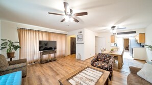 A living room with a flat-screen TV, wooden flooring, a ceiling fan, and a dining area with a table and chairs.