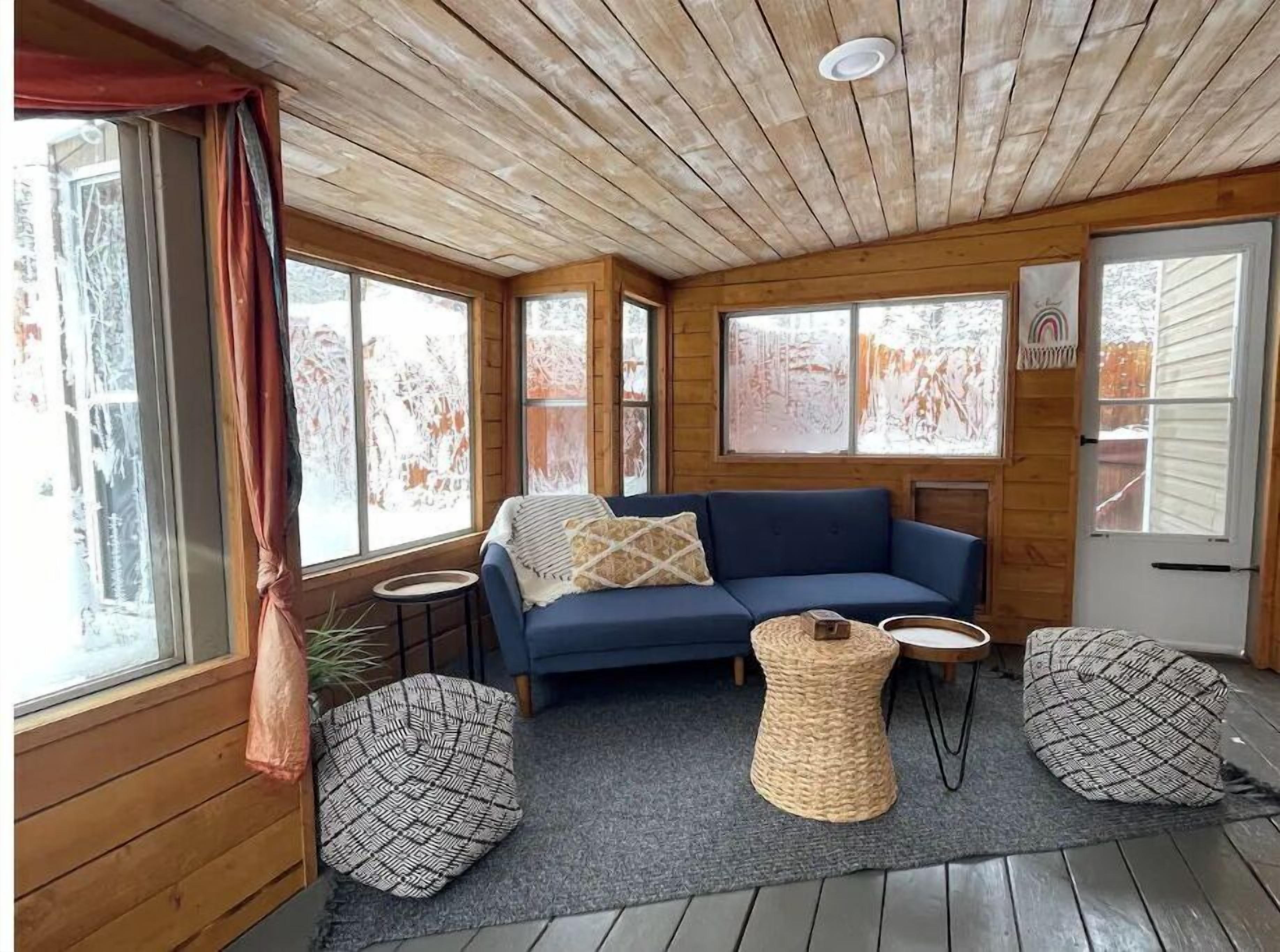 Ein gemütliches Blockhaushaus-Interieur mit einem blauen Sofa, zwei gewebten Poufs, einem kleinen runden Tisch und großen Fenstern mit satiniertem Glas.