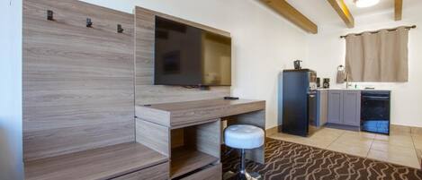 A modern room with a built-in TV, wooden shelving, a small stool, a kitchen area with a refrigerator, and a patterned rug.