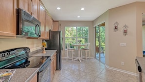 A kitchen with wooden cabinets, a stainless steel refrigerator, a black stove, and a glass-enclosed microwave. There is a window with a view of greenery and a dining area with a table and chairs.