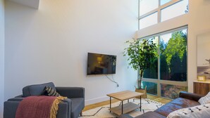 Ein modernes Wohnzimmer mit grauem Sofa, braunem Ledersessel, Couchtisch aus Holz und einem großen Fenster mit Blick auf Grünflächen.