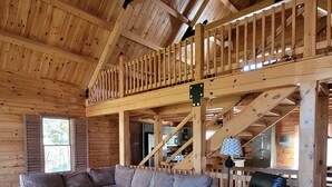 A cozy cabin interior with a wooden staircase, a sectional sofa, a leather armchair, and a patterned rug. - Authentic Log Cabin Getaway w/Private Hot Tub (Berkeley Springs)