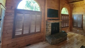A log cabin interior with a fireplace, wooden walls, and large windows. - Authentic Log Cabin Getaway w/Private Hot Tub (Berkeley Springs)