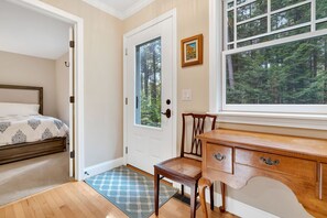 A bedroom with a bed, a wooden dresser, a chair, a door, and a window with a forest view. - Fairbanks Haven House with Hot Tub & Infrared Sauna (Quechee)