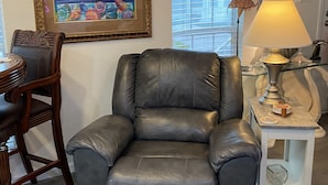 Una sala de estar con un sillón de cuero negro, una mesa de comedor con sillas, un cuadro enmarcado, una lámpara y una ventana con persianas.