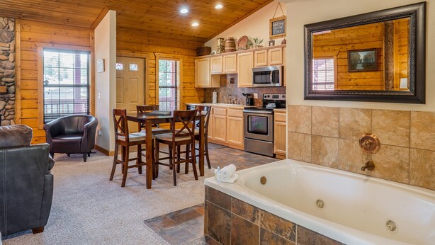 A cozy cabin interior with a stone wall, wooden ceiling, a dining area with chairs, a kitchen with cabinets and appliances, and a large bathtub. - Cozy-up in a Cabin for Two! (Branson)