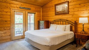 A wooden cabin room with a bed, a nightstand, a lamp, a dresser, and a door with a sign. - Cozy-up in a Cabin for Two! (Branson)