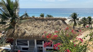 Una cabaña con techo de paja y vista al océano, rodeada de palmeras y buganvillas rosadas.