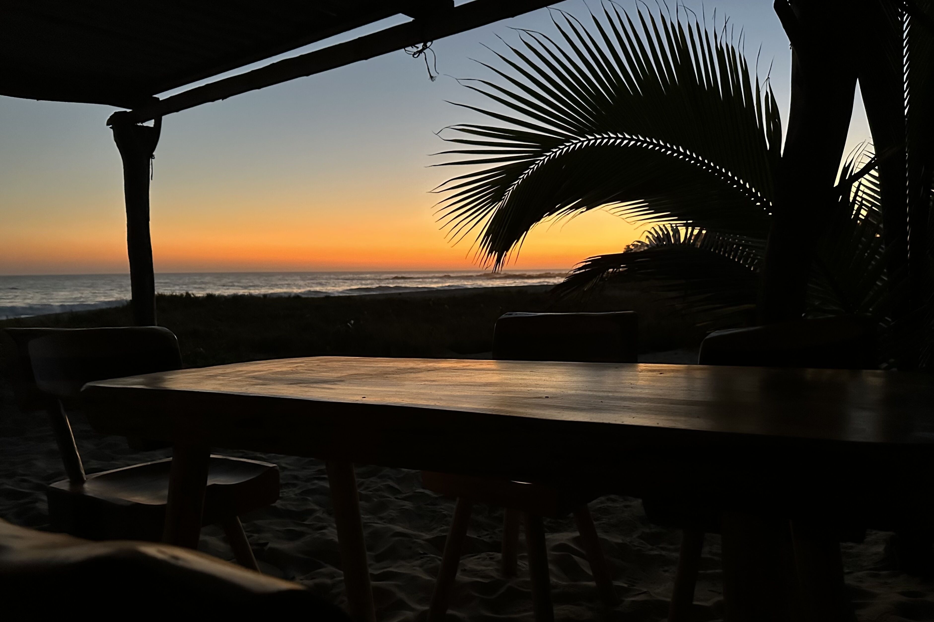 A beachside table and chairs under a shaded area with a view of the ocean at sunset.