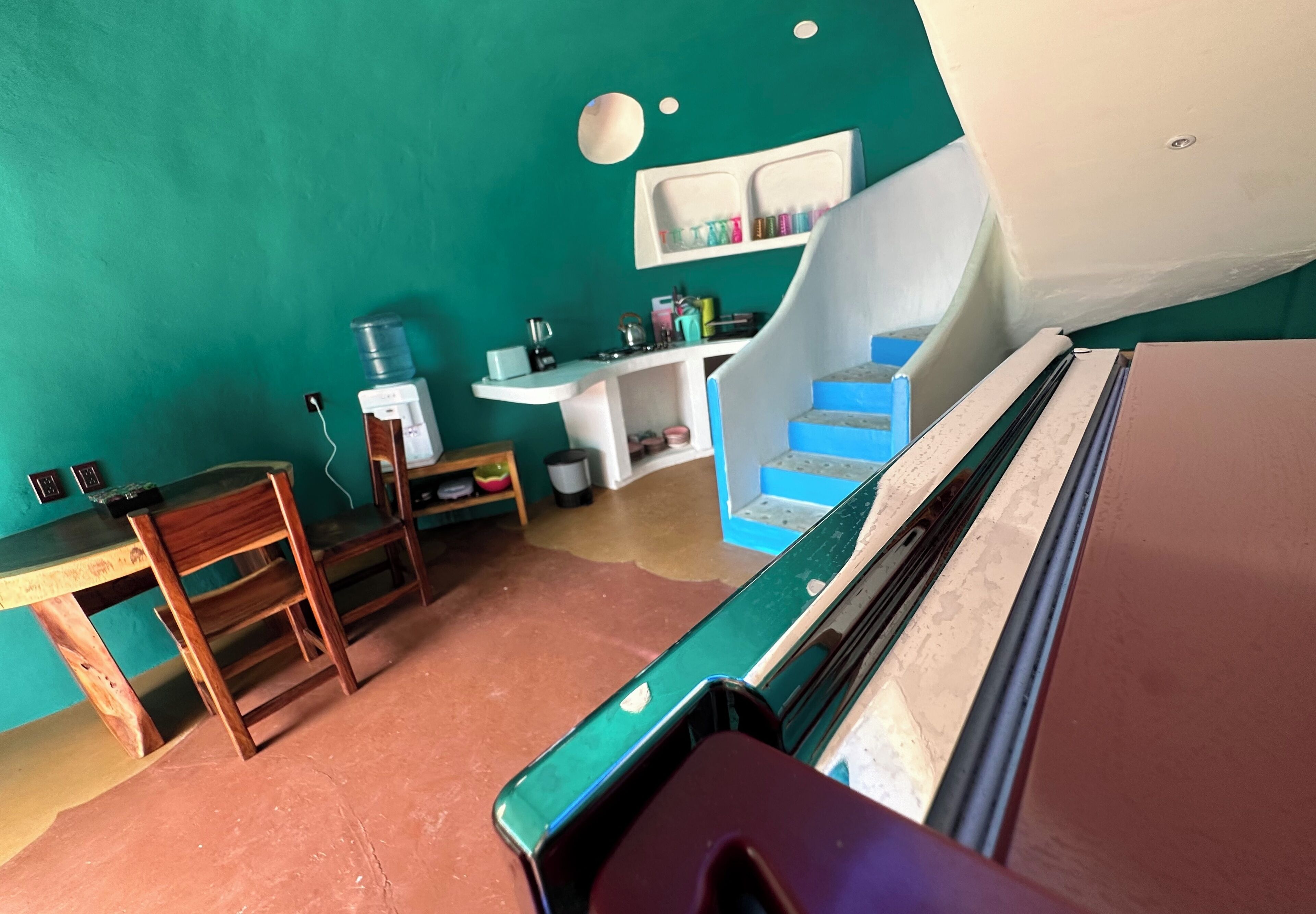 Ein Zimmer mit Türkiswänden, einem Holztisch, einem Stuhl, einem Waschbecken und einer Treppe.