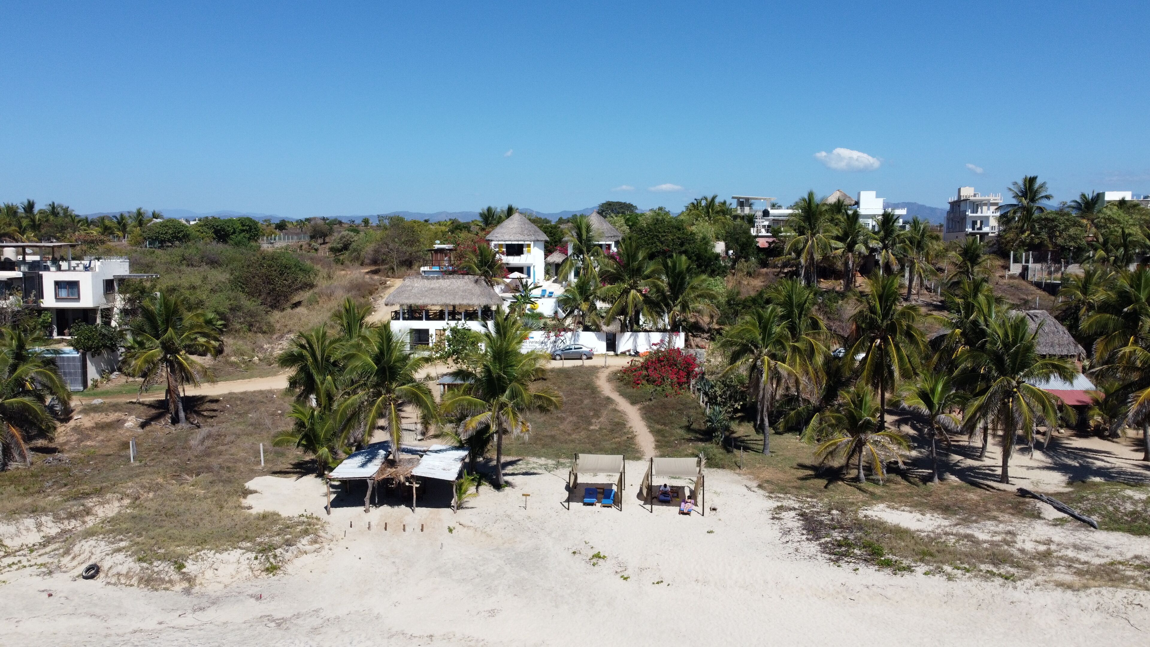 Una propiedad costera con playa de arena, palmeras y un edificio con techo de paja.