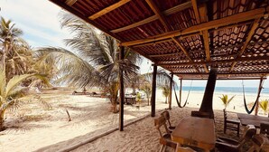 Una cabaña frente a la playa con una mesa y sillas de madera, palmeras y vistas al océano.