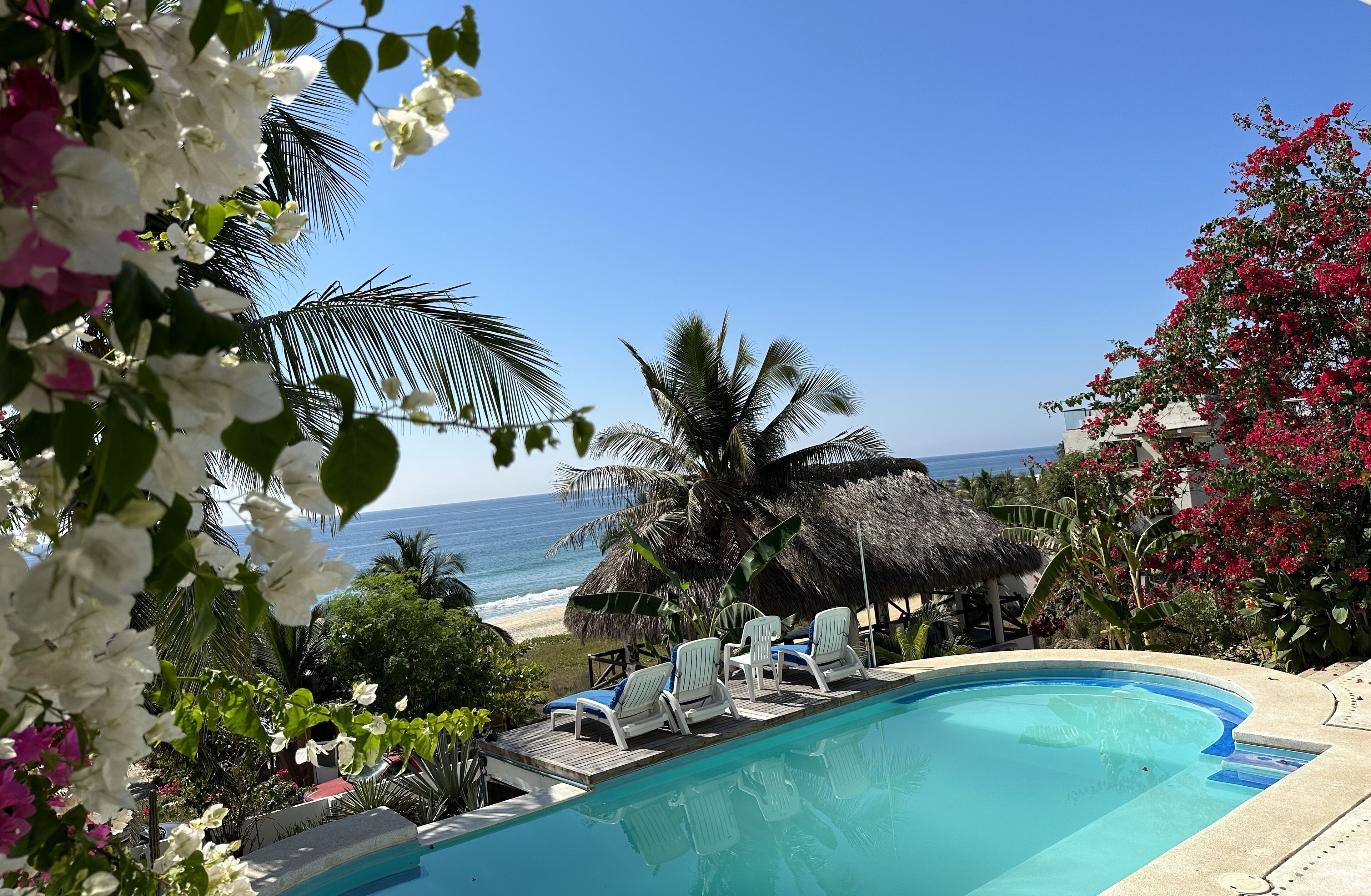 A poolside view with lounge chairs, a thatched-roof structure, and a beach in the background.