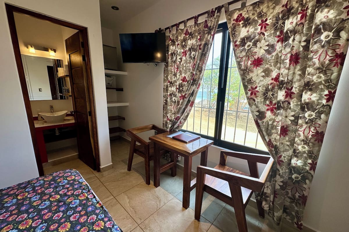 Quarto com cama, escrivaninha com cadeira, televisão na parede, janela com cortinas florais e banheiro com pia e espelho.
