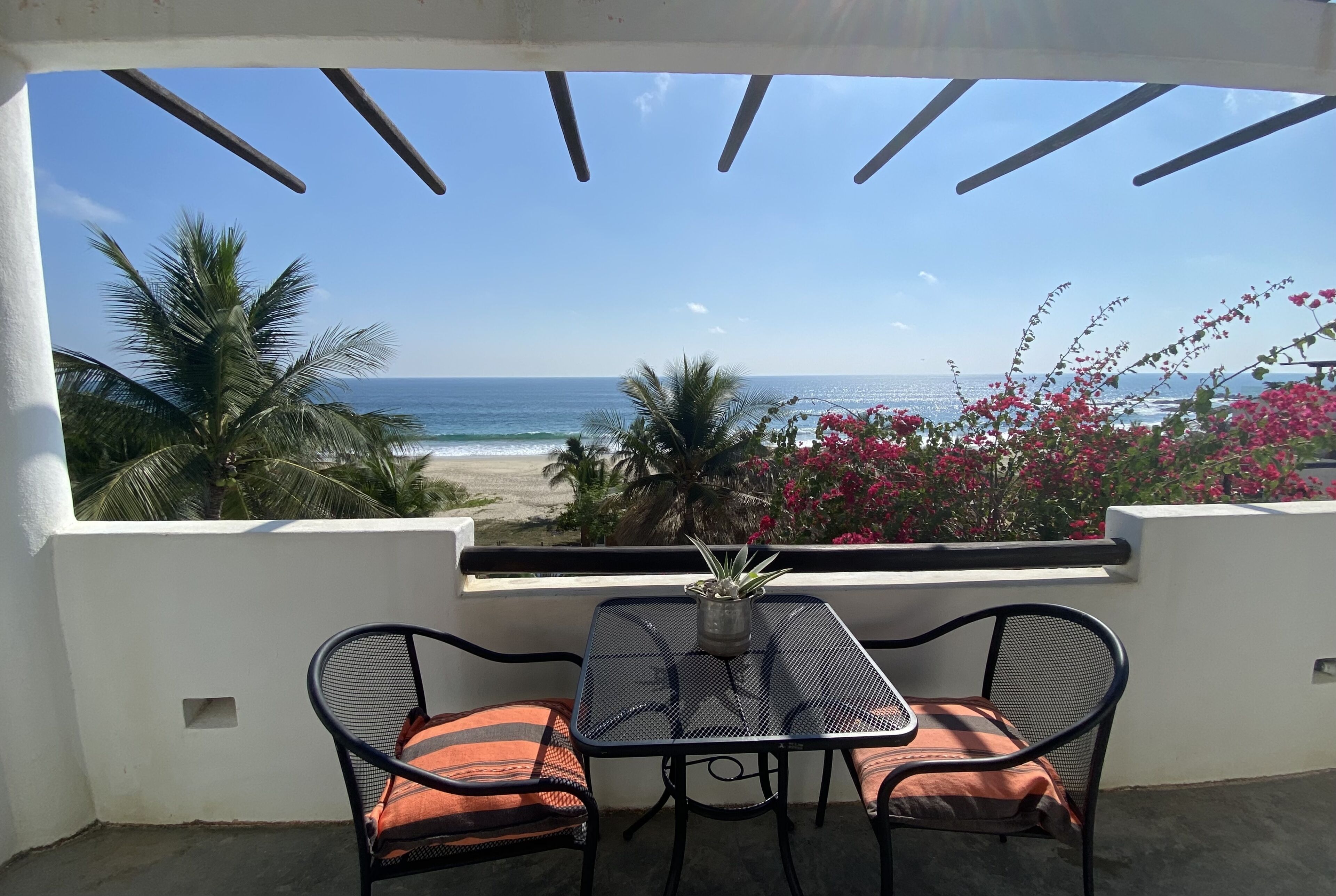 Varanda com mesa e cadeiras, vista para uma praia e palmeiras.