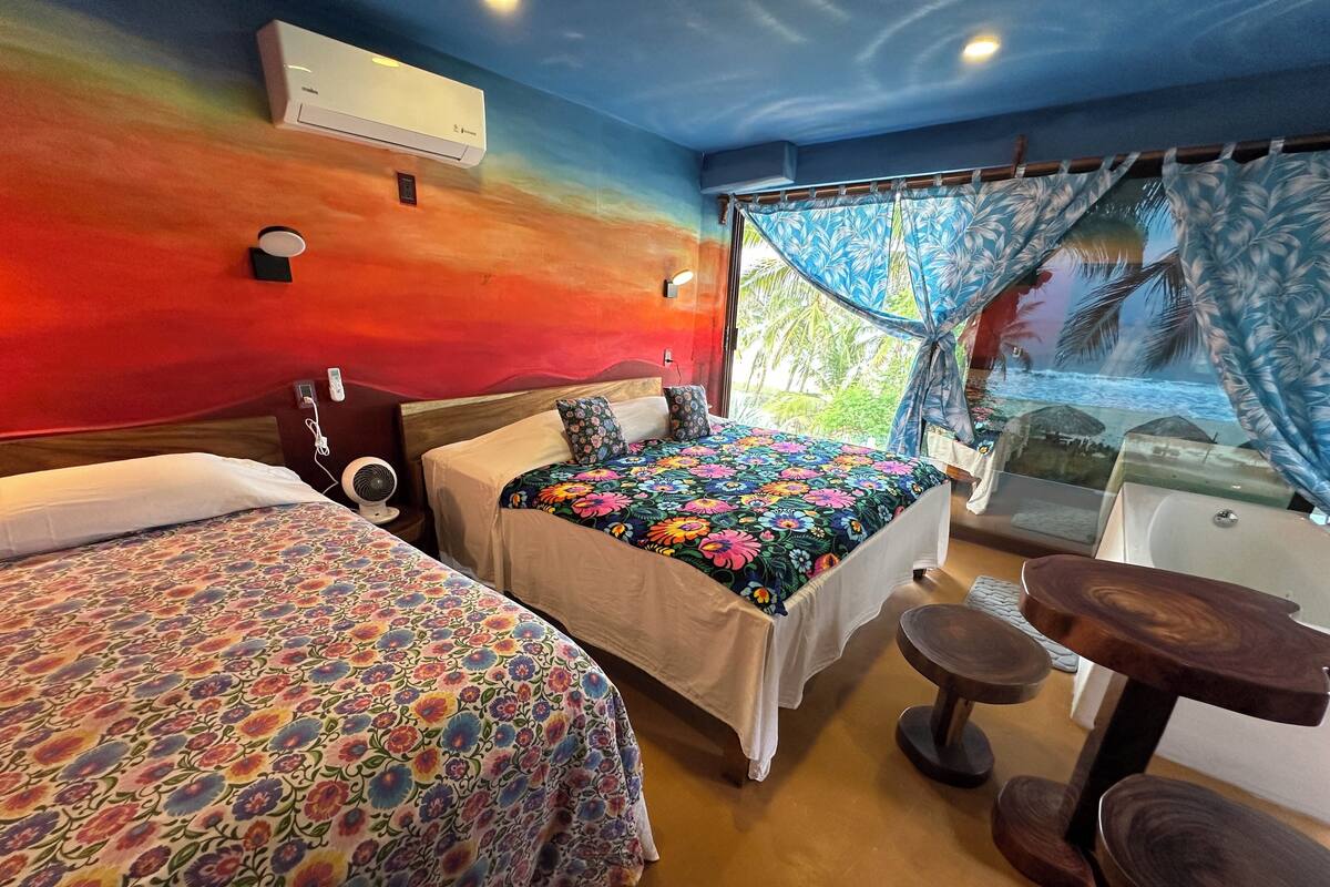 Quarto de hotel com duas camas, uma mesa pequena, um ventilador e uma janela com vista para palmeiras.