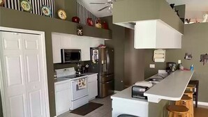 A compact kitchen with white cabinets, a microwave, a stove, and a refrigerator. There is a trash bin and wooden stools near the counter. - Cheery 4 Bedroom Pool Villa (Valrico)