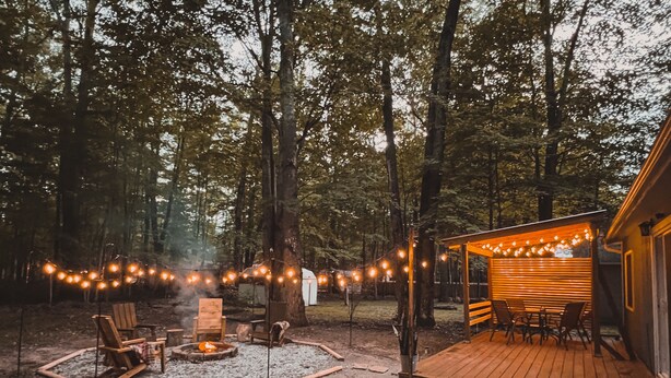 Un espace extérieur aménagé dans un style rustique, avec un foyer, une terrasse en bois et des guirlandes lumineuses.