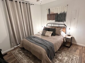 Une chambre à coucher comprenant un lit, une table de chevet, une lampe, une décoration murale en macramé et une fenêtre avec des rideaux.