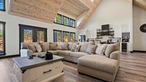 Un vaste salon comprenant un grand canapé d’angle, une table basse et une commode intégrée. De grandes fenêtres laissent entrer la lumière naturelle et offrent une vue sur l’extérieur.