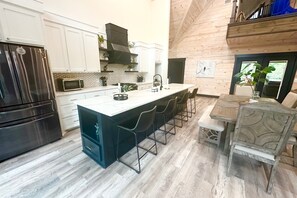 A modern kitchen with a large island, bar stools, a dining table with chairs, and a view of the outdoors. - Swim Spa | Indoor Slide | Hot Tub | Disc Golf (Broken Bow)