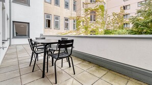 An outdoor patio with a table and chairs, a tiled floor, and a view of adjacent buildings with windows and greenery.