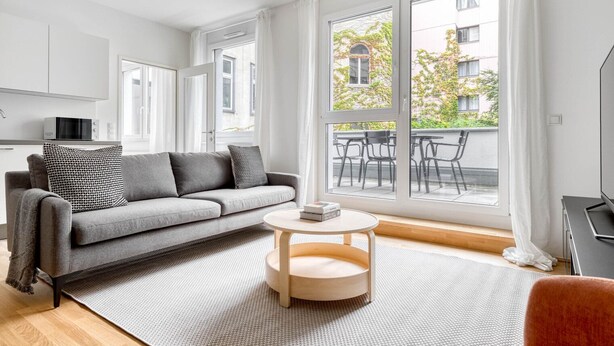 A modern living room with a grey sofa, a round wooden coffee table, and a large window with a view of the outside.