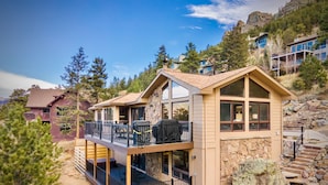 A two-story cabin with a balcony, stone foundation, and a view of surrounding mountains.