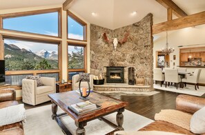 Relax in a spacious living area with a high ceiling and stunning mountain views, featuring a stone fireplace and comfortable leather sofas.