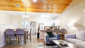 A spacious living room with a wooden ceiling, a dining area with a wooden table and chairs, a kitchen with white cabinets, and a gray sofa. - Shared Heated Pool With Slide, Golf Cart Accessible, Pet Friendly (Port Aransas)