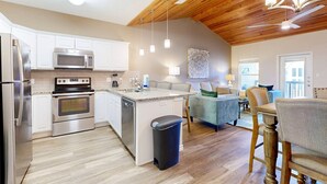 A modern kitchen with stainless steel appliances, a wooden ceiling, and a dining area with a blue sofa and wooden chairs. - Shared Heated Pool With Slide, Golf Cart Accessible, Pet Friendly (Port Aransas)