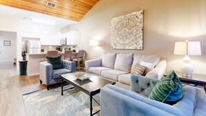 A modern living room with a wooden ceiling, a large abstract painting, and a kitchen in the background. - Shared Heated Pool With Slide, Golf Cart Accessible, Pet Friendly (Port Aransas)