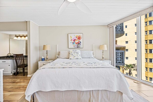 A bedroom with a bed, bedside lamps, a painting, and a view of buildings. - Oceanfront Condo with Private Balcony Heated Pool and Walk to Flagler Avenue (New Smyrna Beach)