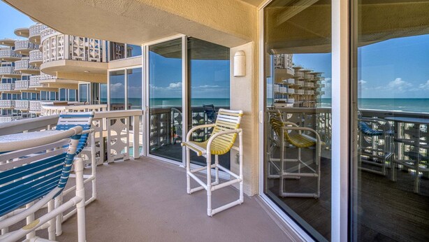 A balcony with a view of the ocean, white outdoor furniture, and a glass door leading to another area.