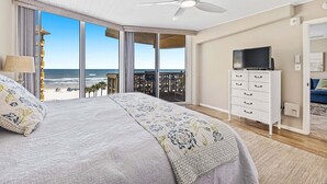 A bedroom with a bed, a ceiling fan, a TV on a dresser, and a view of the beach.