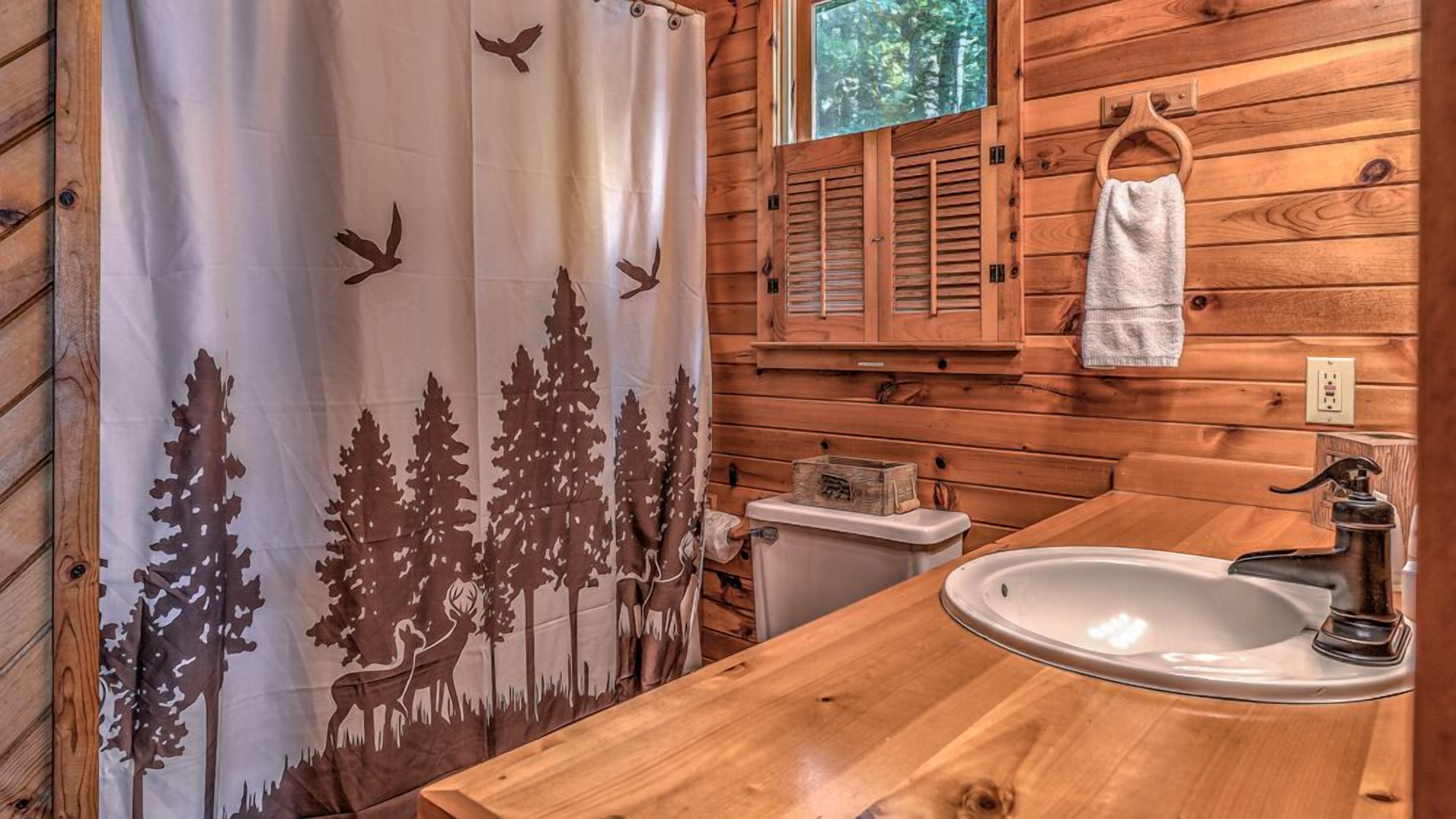 A wooden cabin bathroom with a shower curtain featuring forest and bird silhouettes, a wooden sink, and a window with shutters.