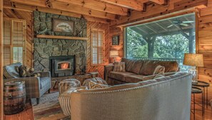 A cozy cabin interior with a stone fireplace, wooden walls, a leather sofa, and a large window overlooking greenery.