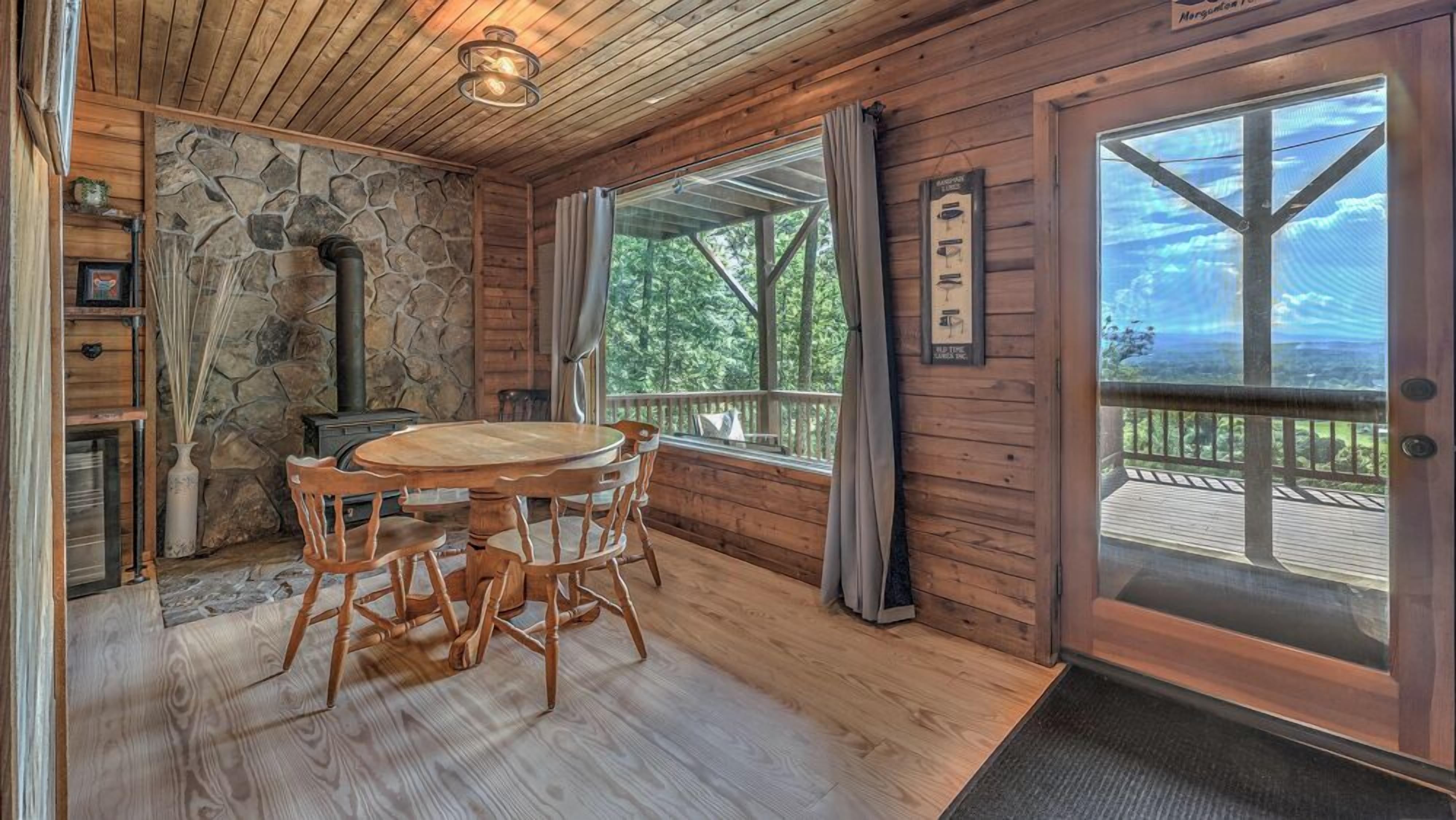 L’intérieur d’un chalet en bois, avec un mur en pierre, une cheminée, une table ronde en bois entourée de chaises et une grande fenêtre donnant sur l’extérieur.